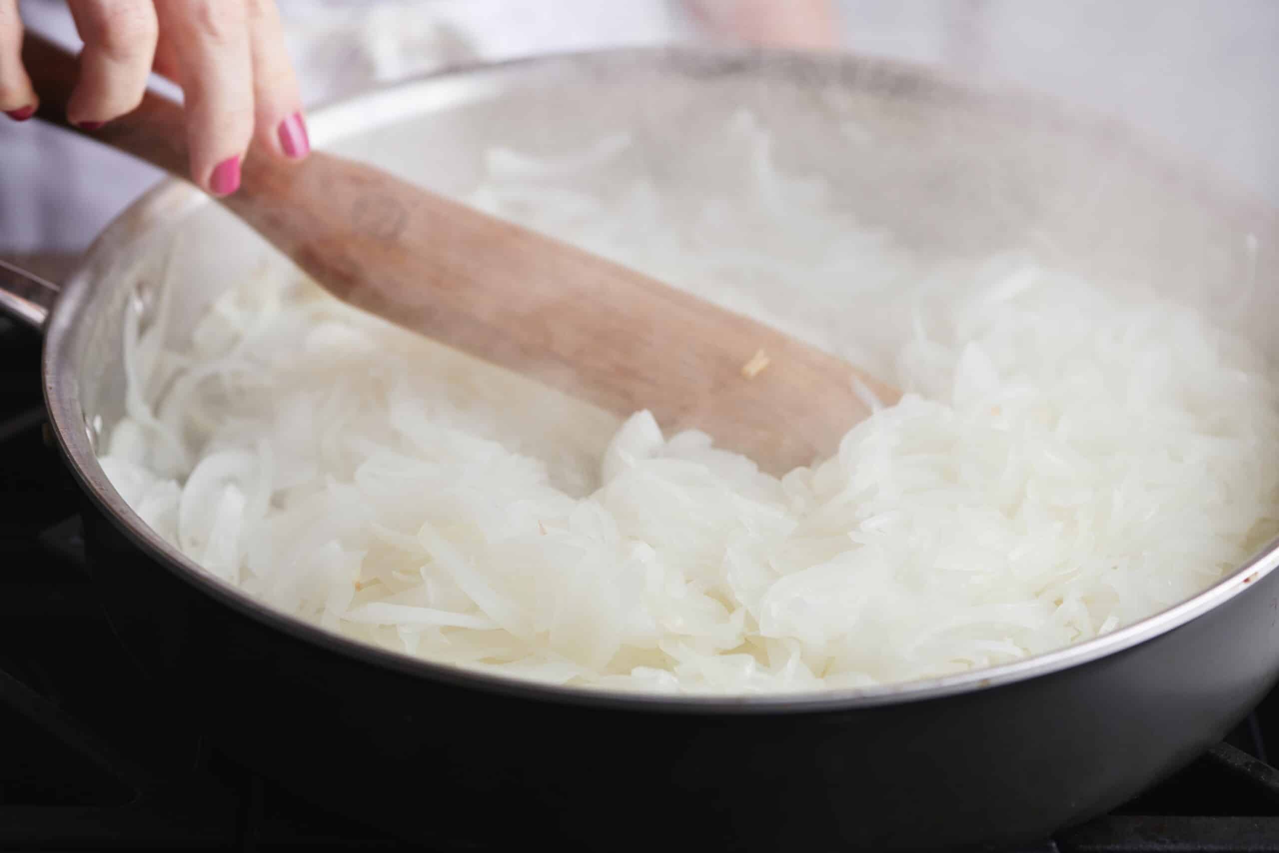 Woman using a wooden spatula on a pan of thinly-sliced onions.