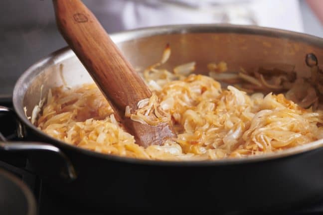 Partially-Caramelized Onions turning brown in a pan with a wooden spatula.