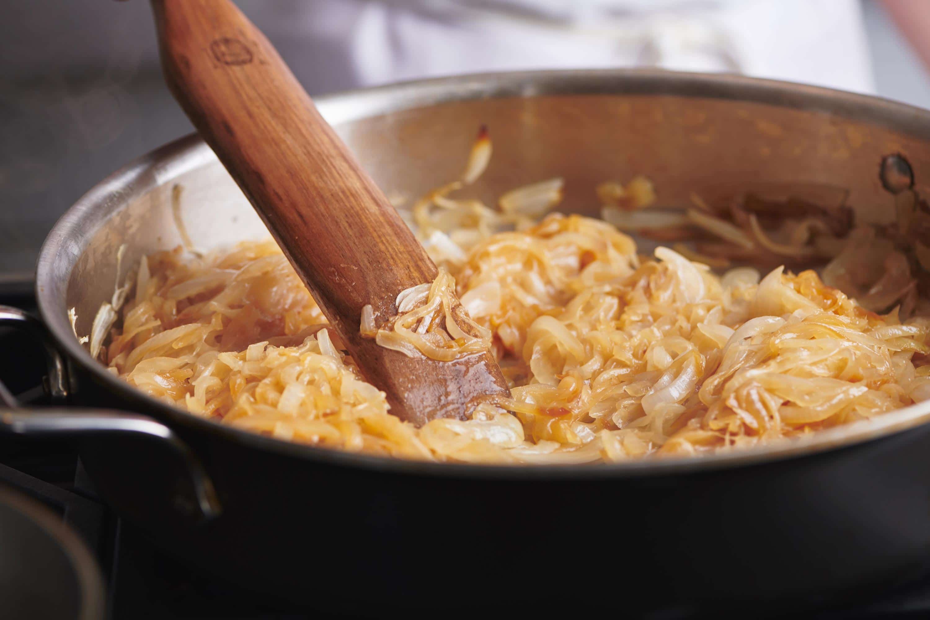 Partially-Caramelized Onions turning brown in a pan with a wooden spatula.