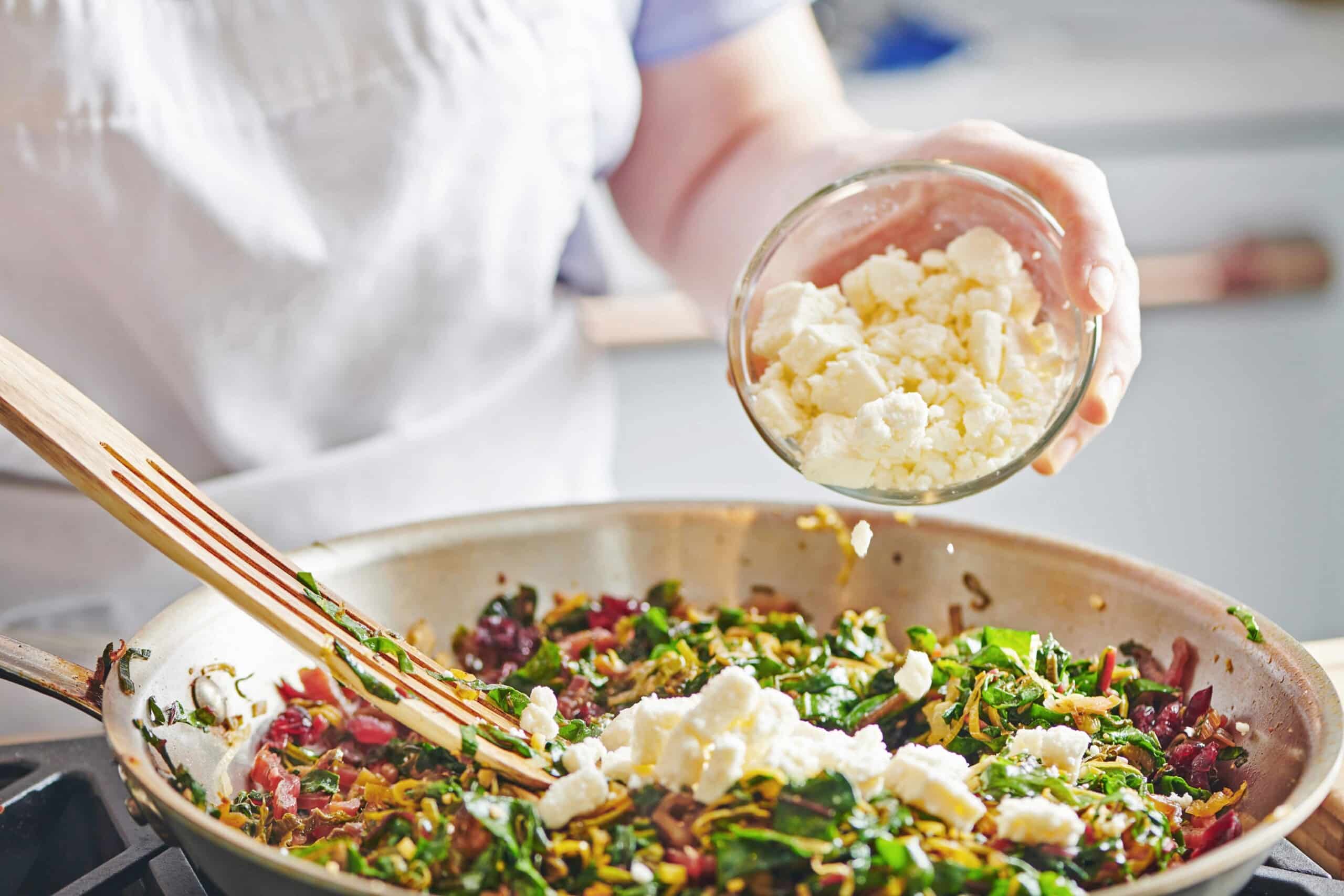 Woman putting feta into a skillet with swiss chard and leeks.