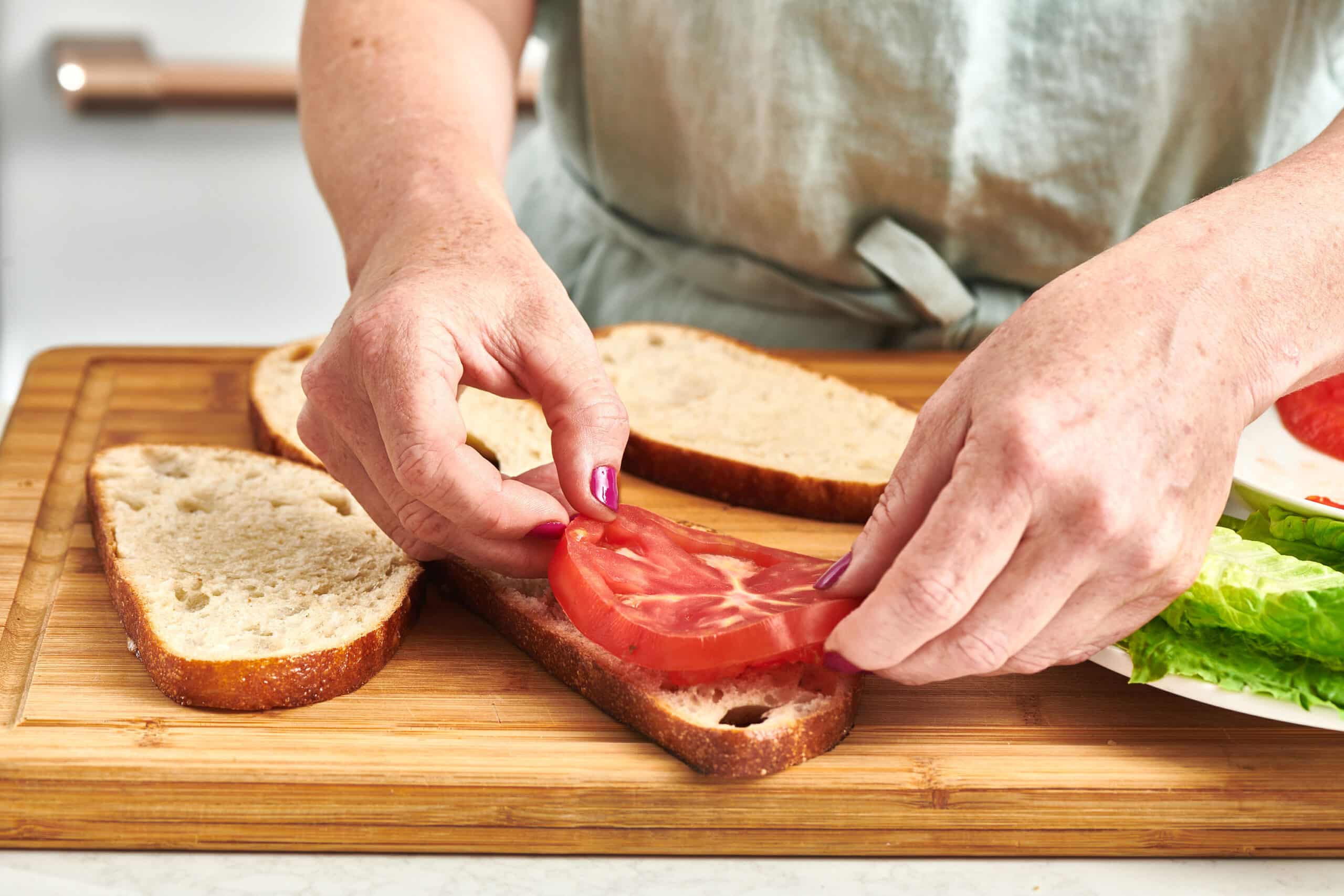A woman's hands placing a slice of red tomato on a piece of bread on a cutting board.
