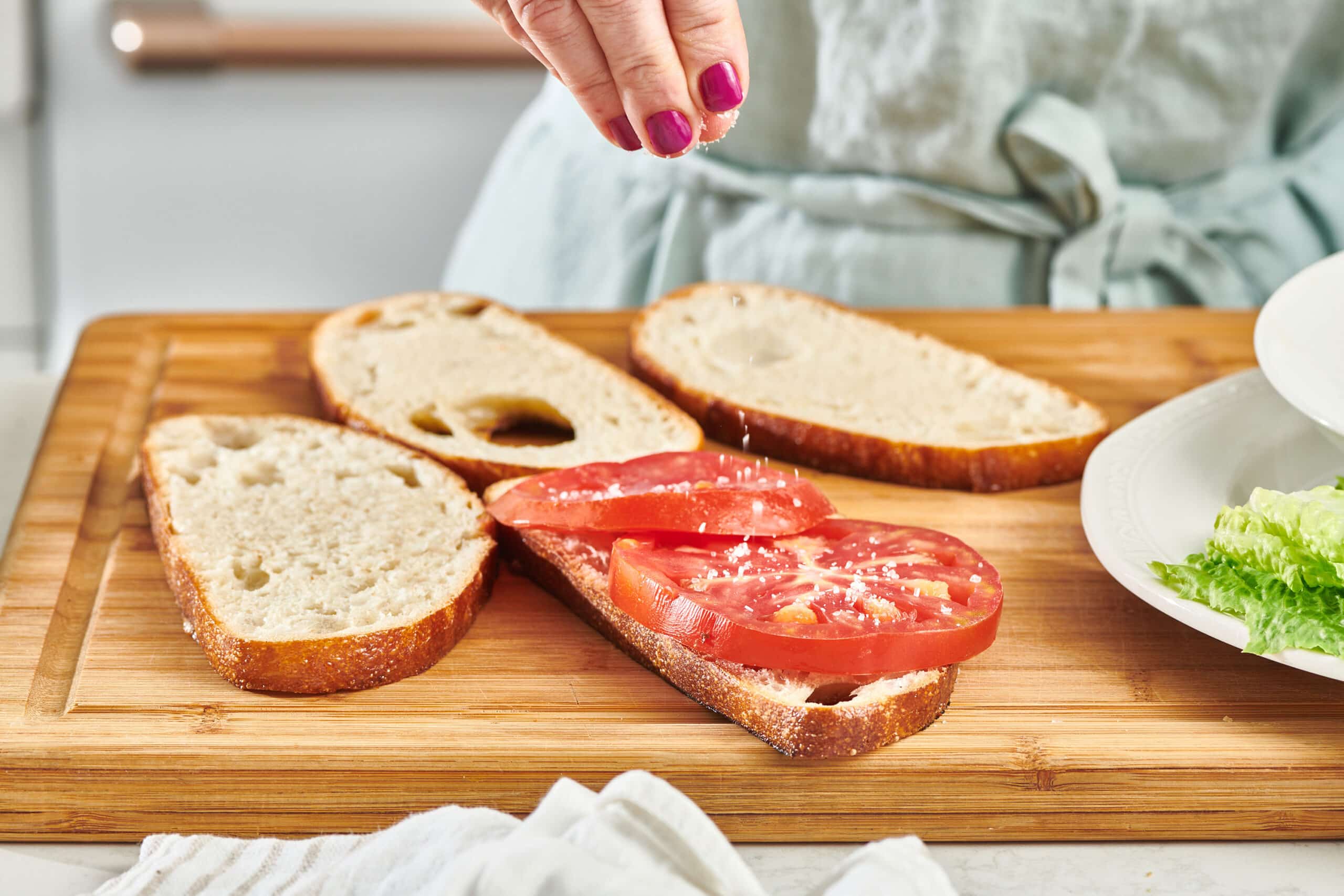 A woman's hand sprinkling kosher salt on a tomato placed on a piece of bread on a cutting board, with other slices of bread on the board.