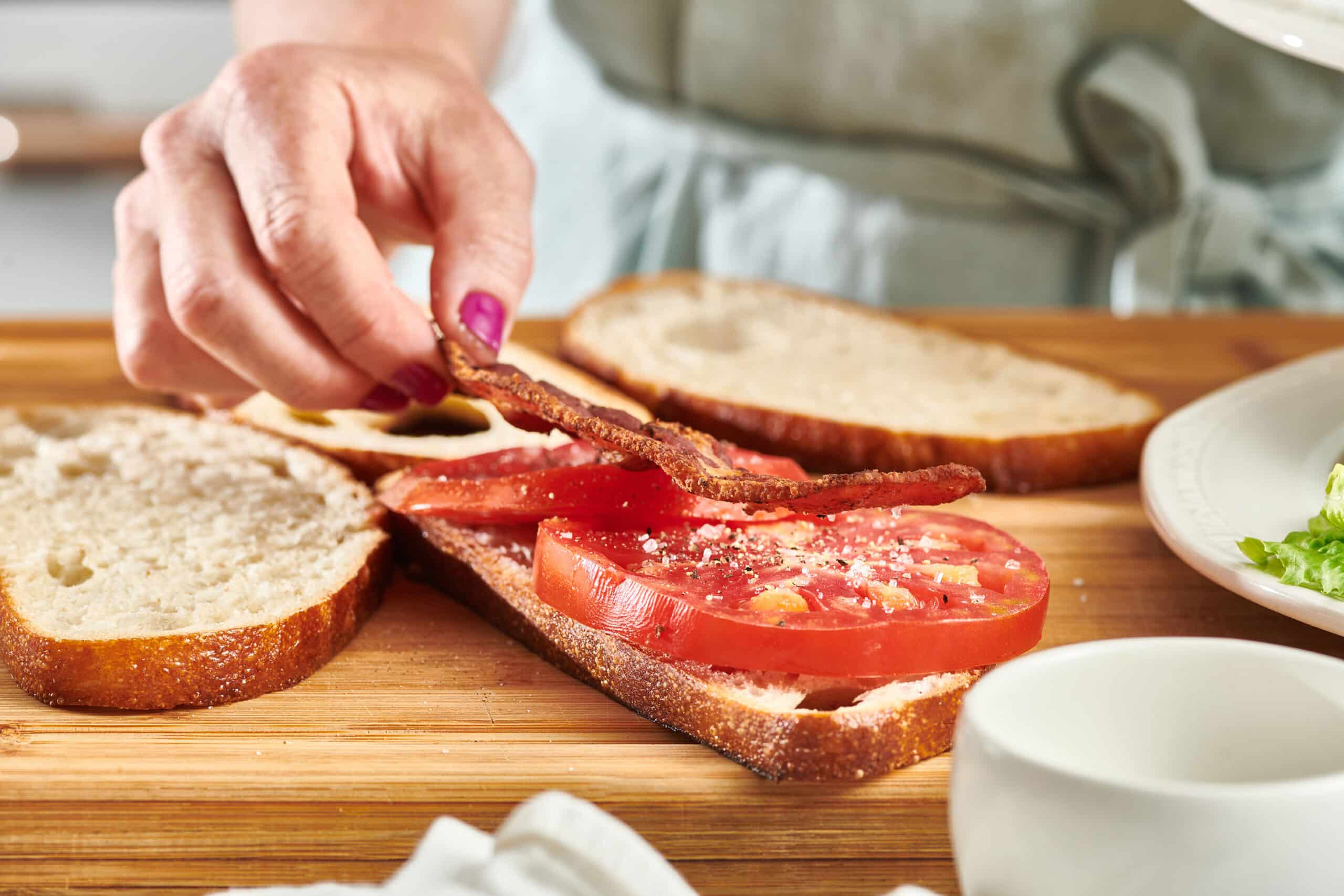 A woman's hand placing a slice of bacon on a slice of tomato resting on a slice of bread on a wooden cutting board.  