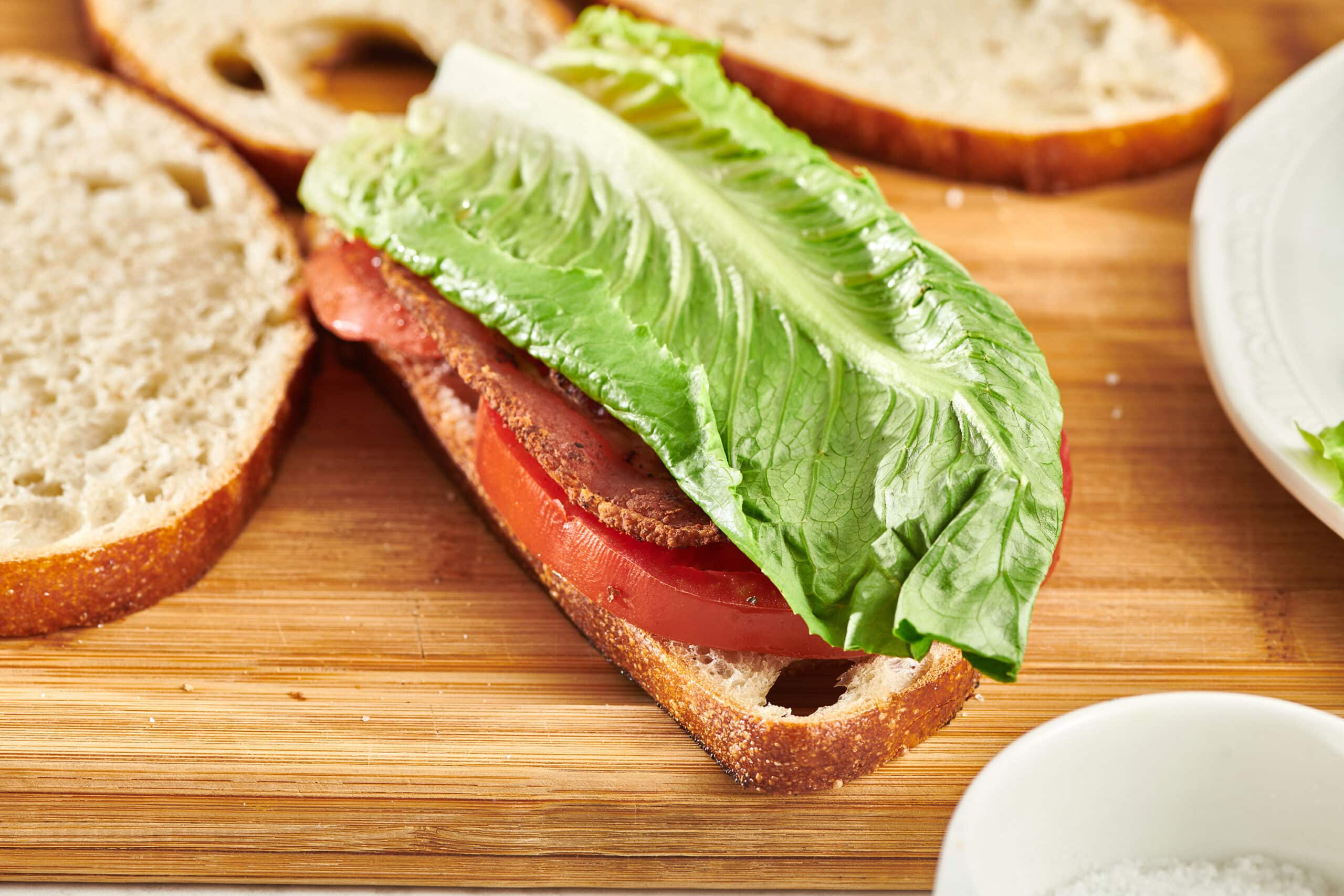 An open sandwich on a wooden cutting board with a piece of lettuce on top of a bacon and sliced tomato on bread.  