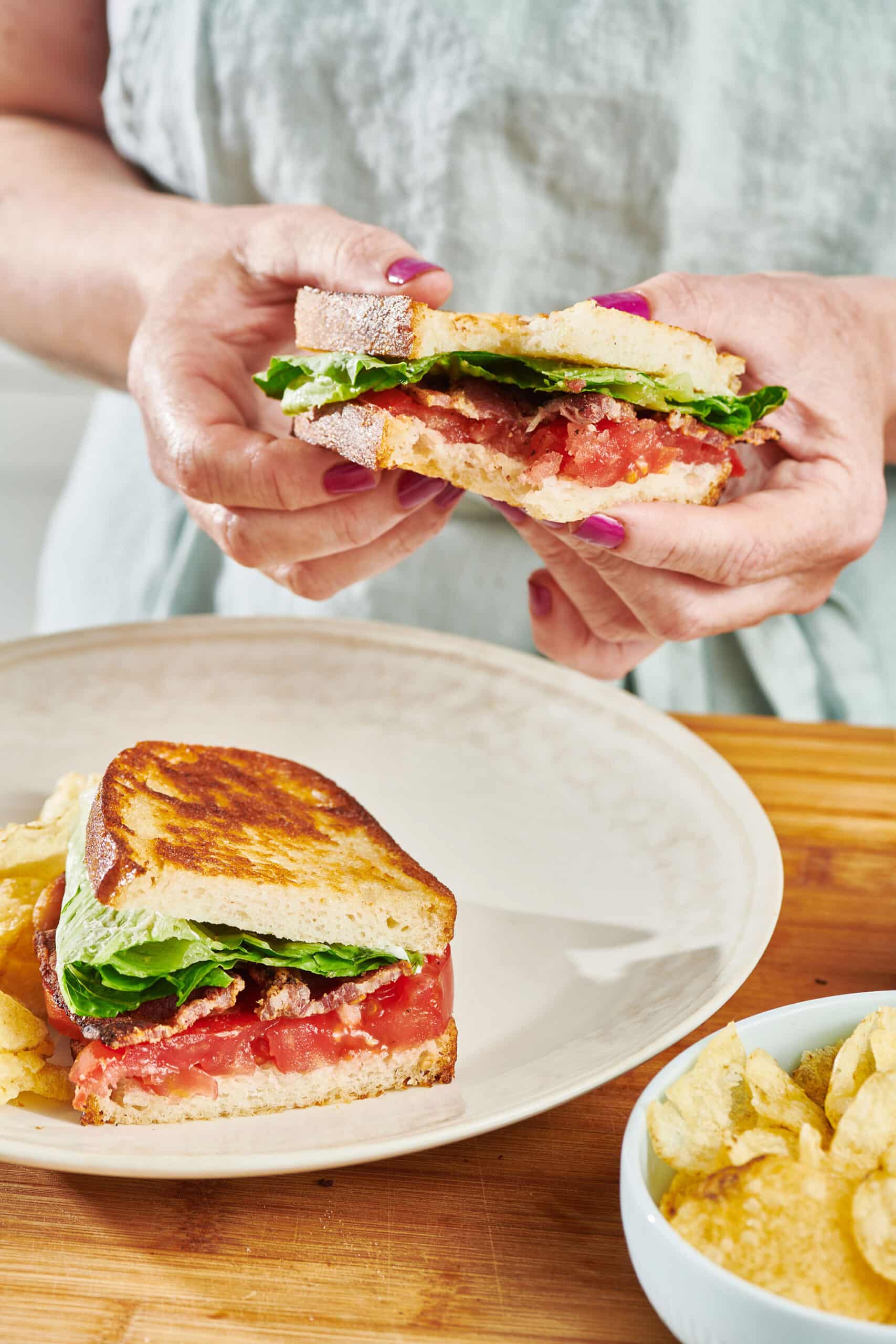 A woman holding a BLT with a bite taken out of it.