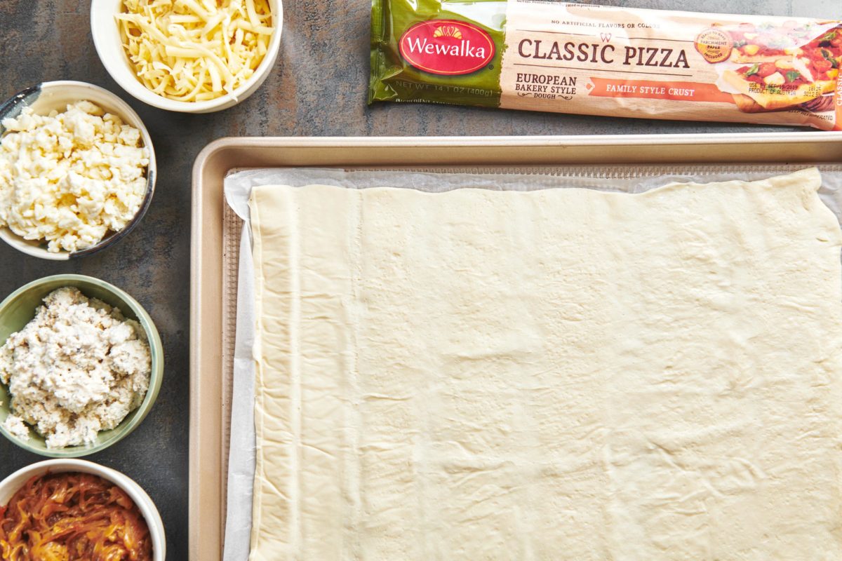 Wewalka pizza dough spread on a baking sheet next to bowls of ingredients.