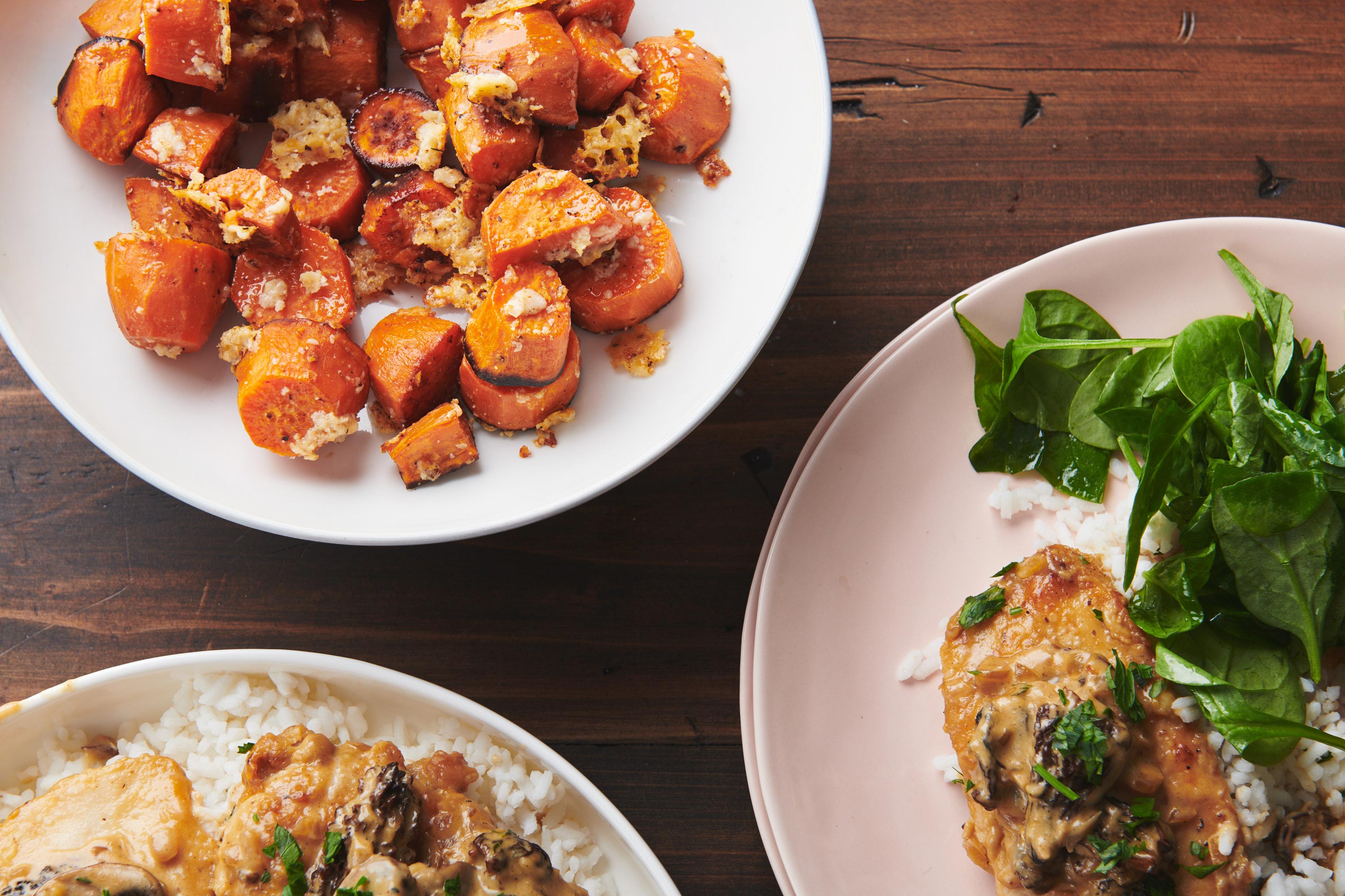 Garlic-Parmesan Sweet Potatoes in a bowl next to plates with meat, rice, and greens.