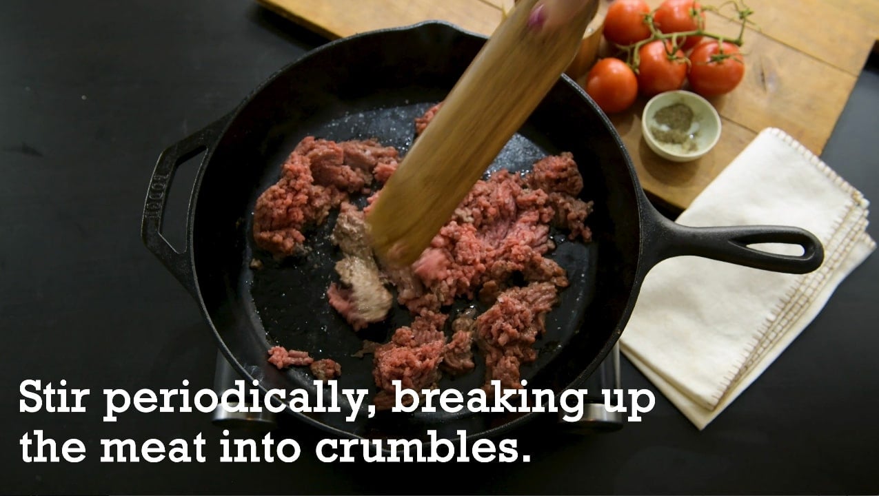 Breaking up ground beef into crumbles as it cooks in cast-iron pan.
