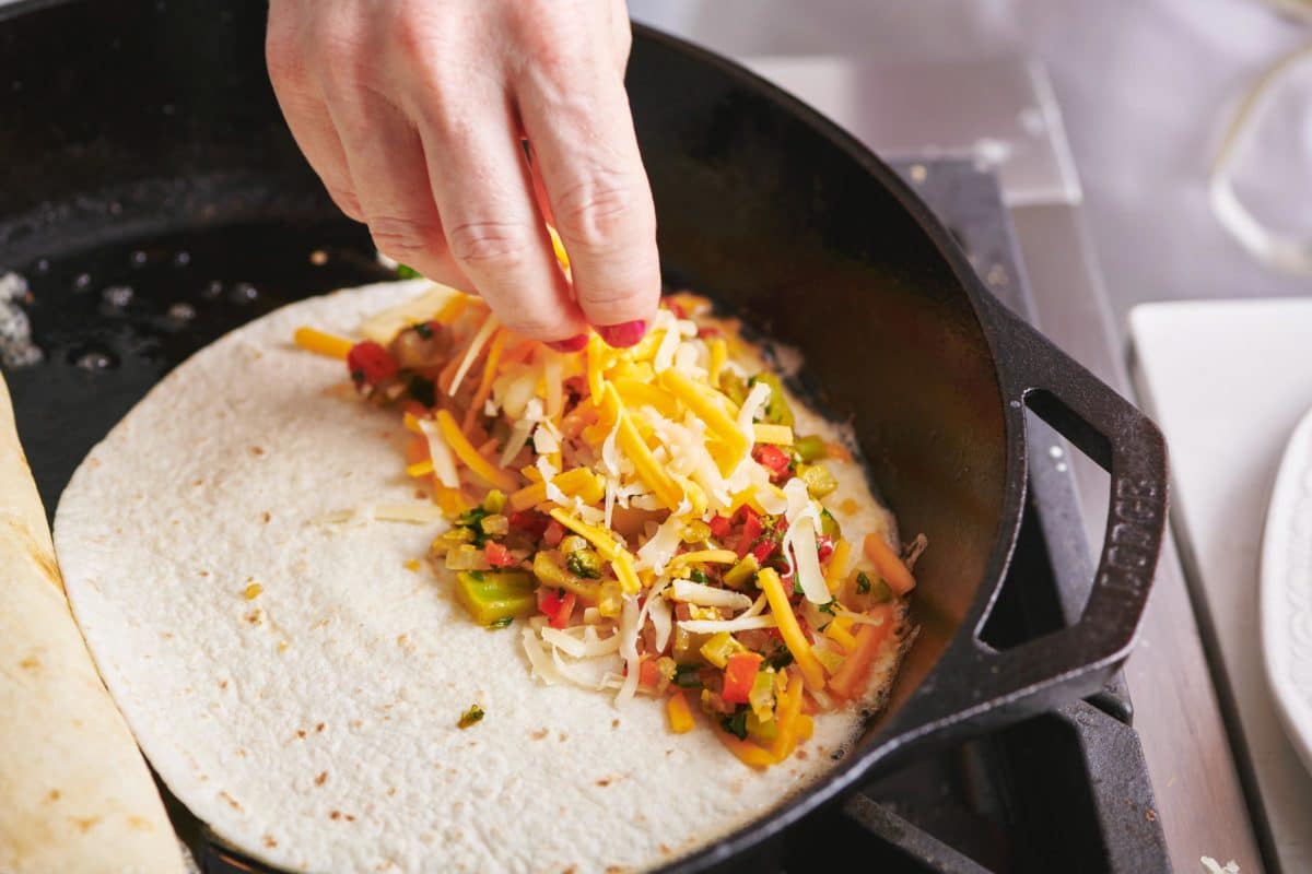 Woman sprinkling more cheese onto a tortilla with cheese and vegetable filling.