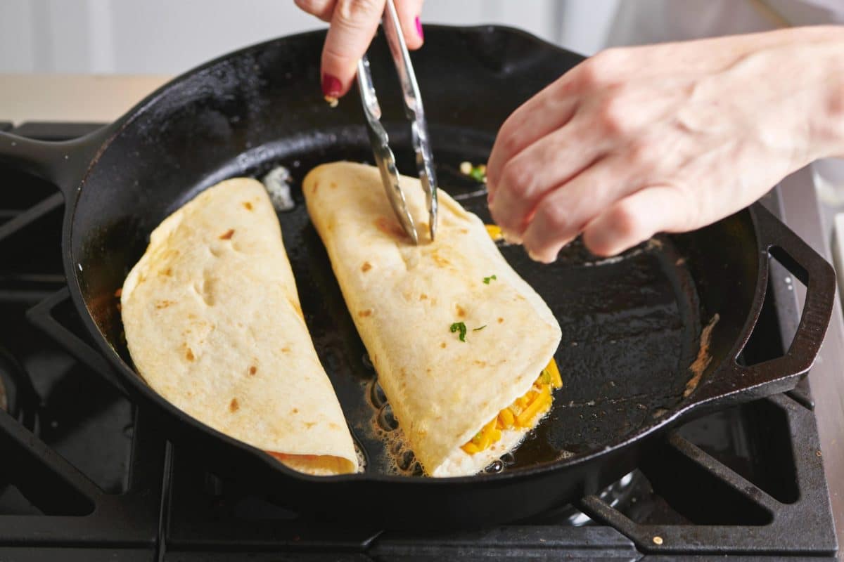 Woman using tongs to fold a tortilla with filling in a skillet.