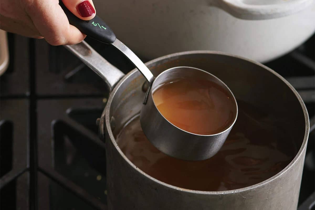 Woman holding a measuring cup of broth over small saucepan.