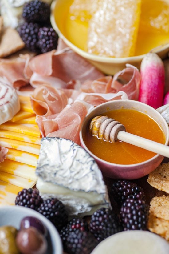 Bowl of honey next to meat, cheese, fruit, and other grazing board foods.