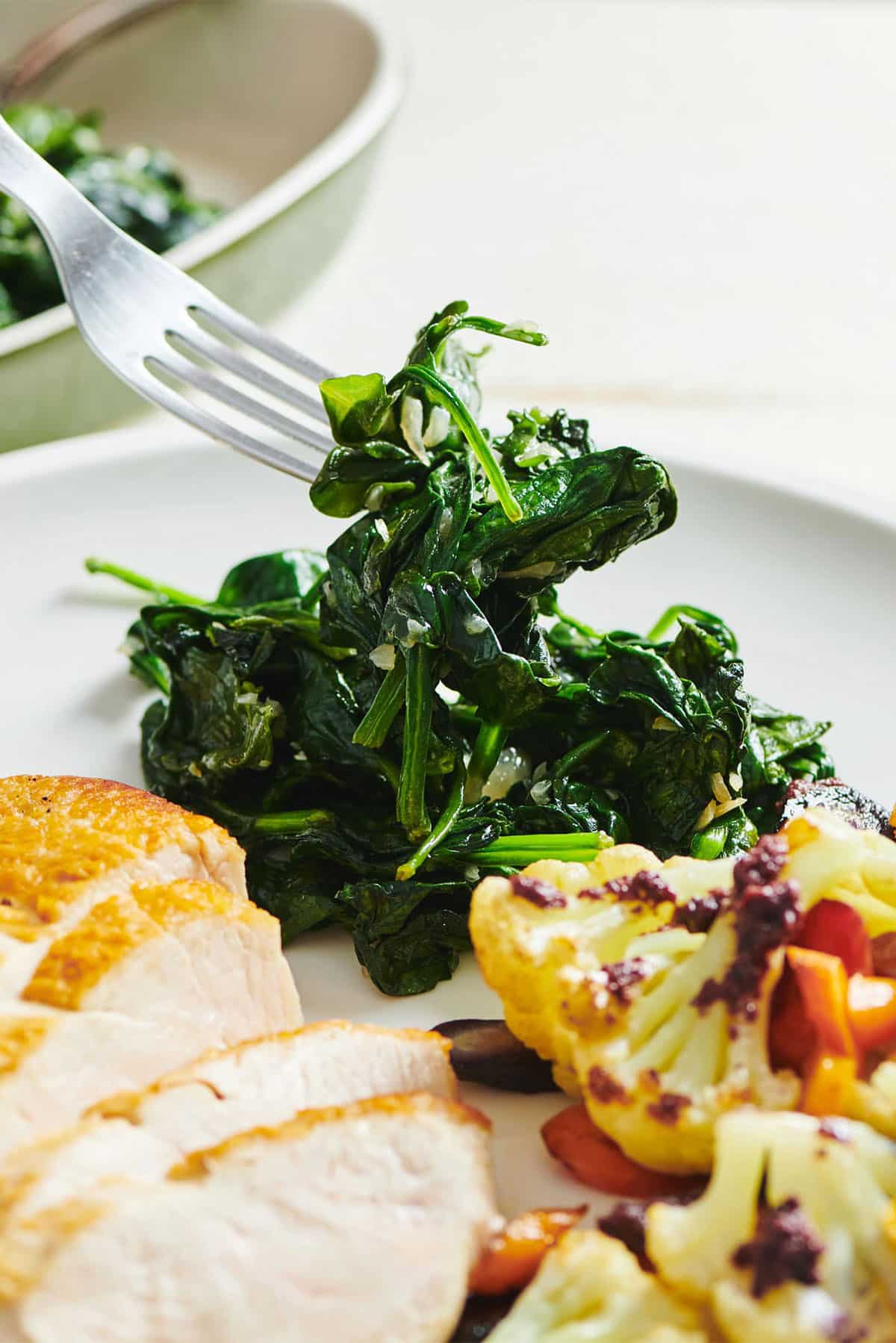 Fork picking up sauteed spinach from plate with chicken and vegetables.