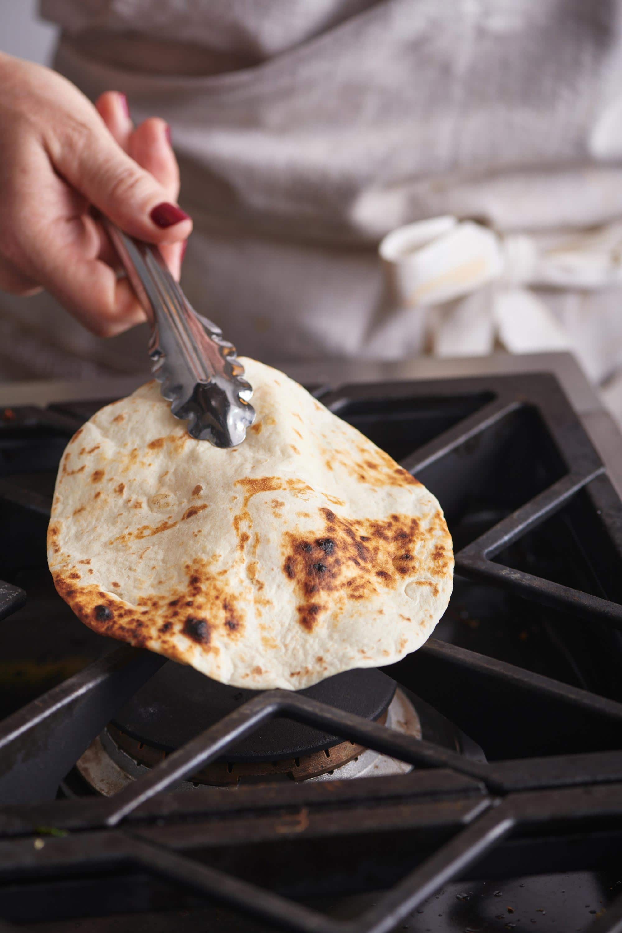 Woman using tongs to flip a tortilla directly on a gas burner.