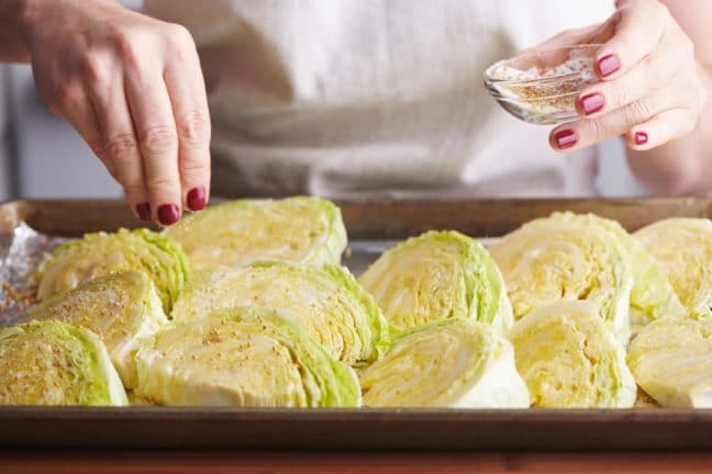 Woman sprinkling salt and mustard seeds onto a baking sheet of cabbage wedges.