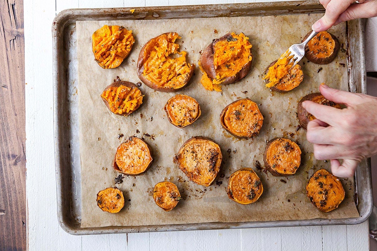 Fork smashing Garlic Butter Sweet Potatoes on a lined baking sheet.