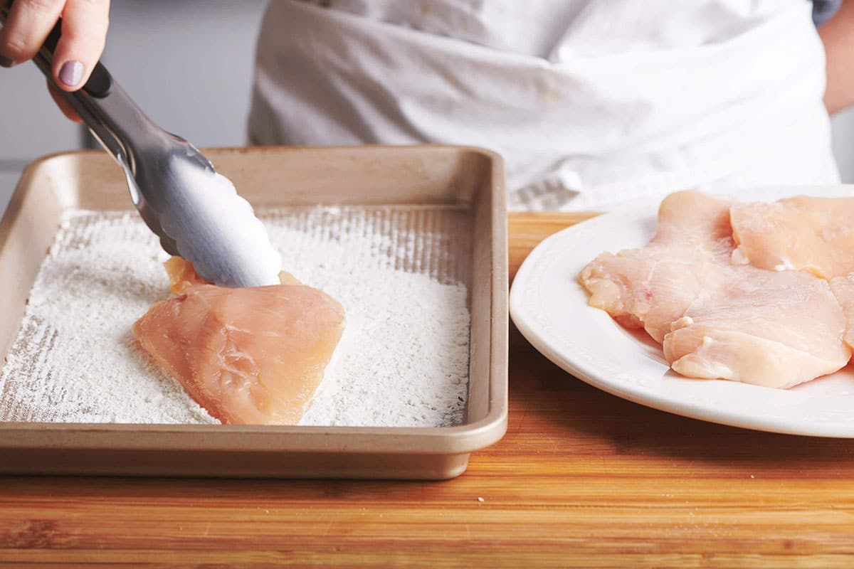 Woman using tongs to dredge chicken in flour.