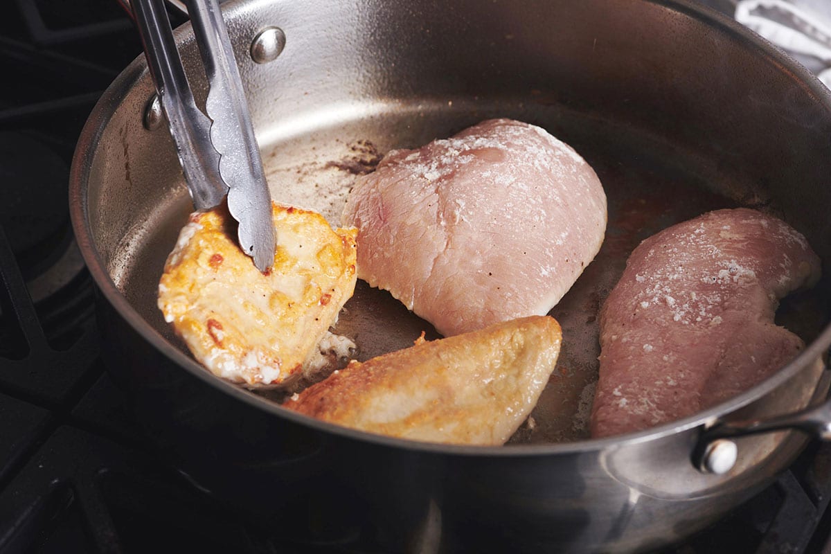 Woman using tongs to flip floured chicken in a skillet.