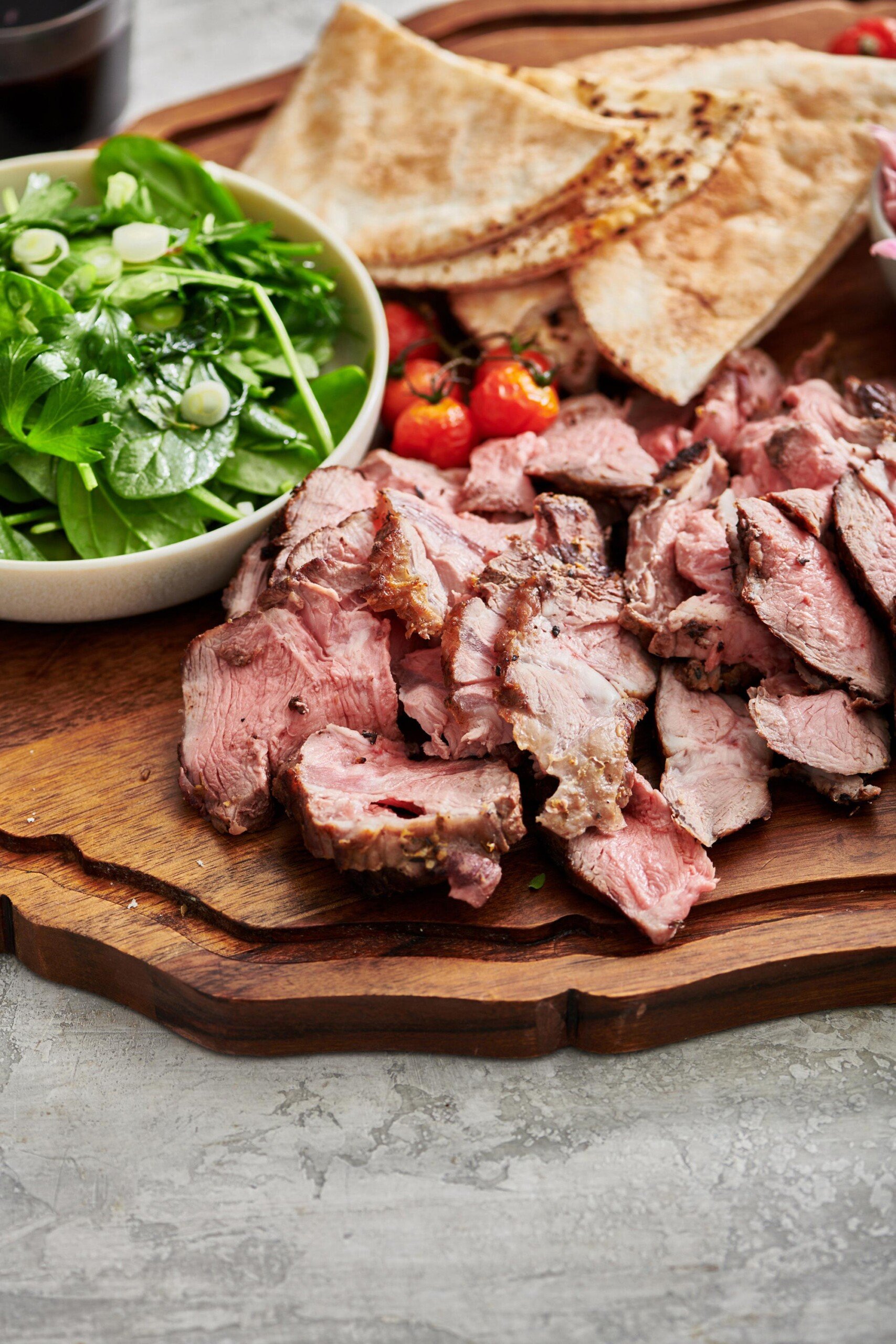 Sliced lamb on a board with pitas and a spinach salad.