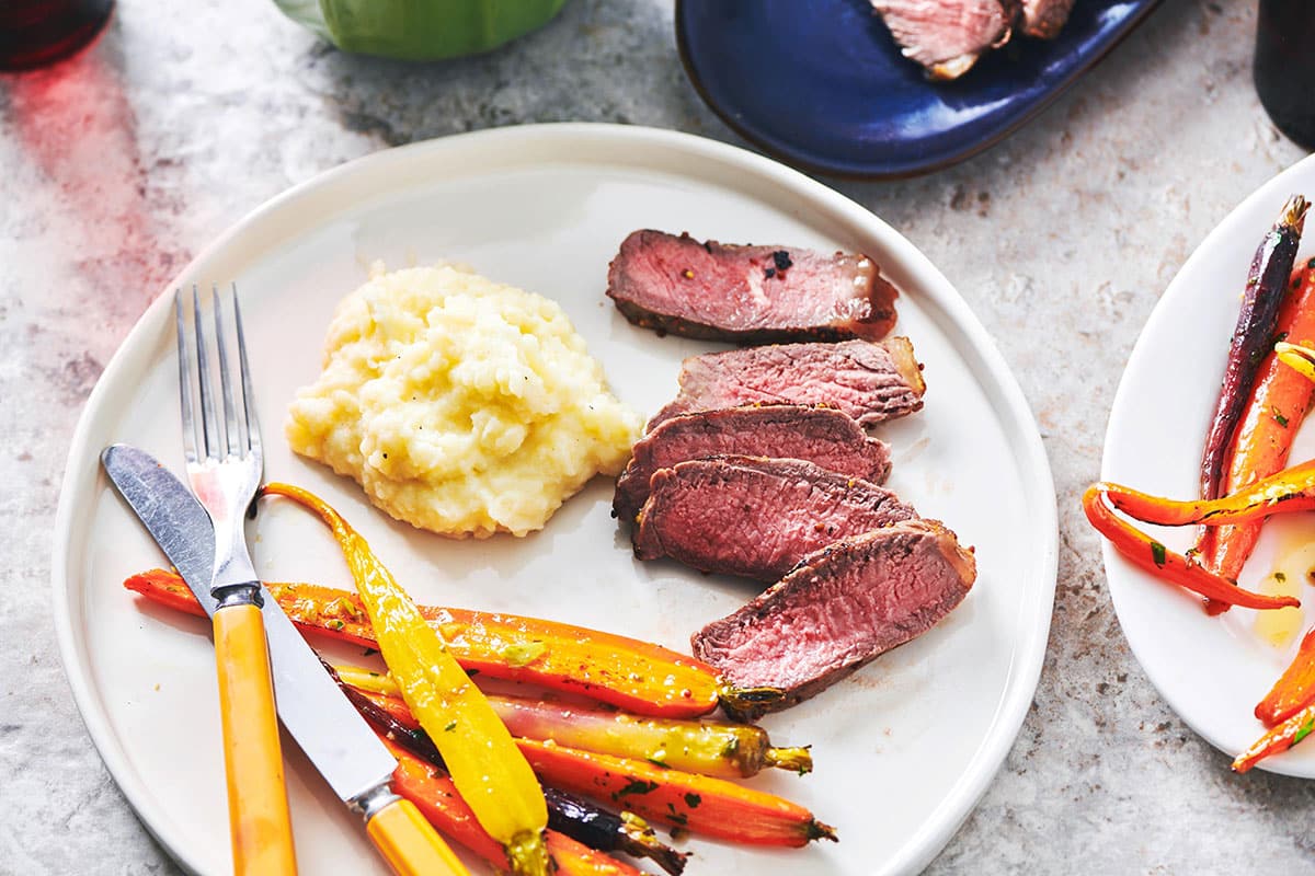Pepper-Mustard Strip Steak sliced on plate with mashed potatoes and roasted carrots.