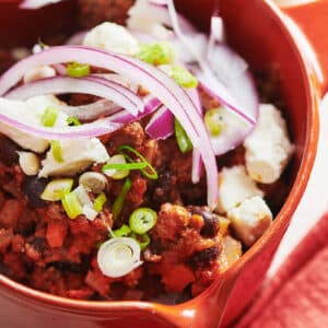 Spicy beef chili with jalapeno in red bowl with toppings.