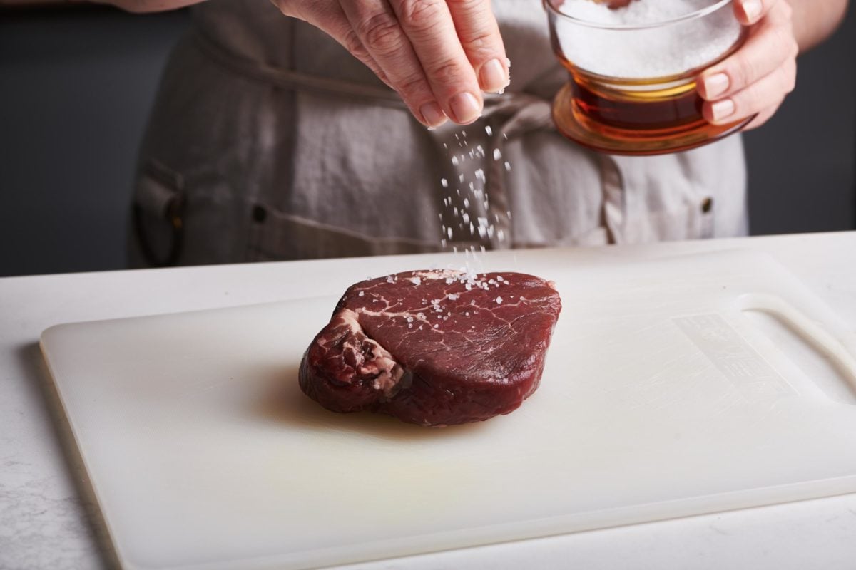 Woman sprinkling salt onto an uncooked filet mignon.