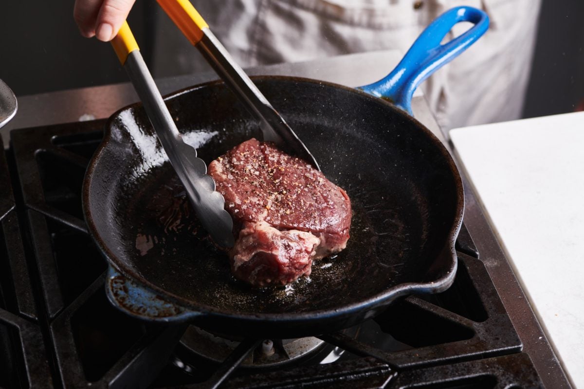 Tongs grabbing a Filet Mignon in a cast iron skillet.