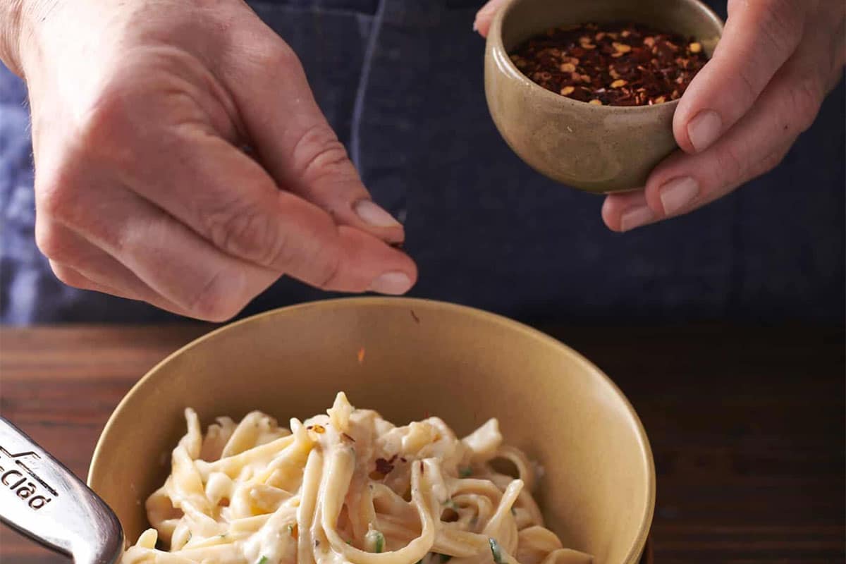 Sprinkling red pepper flakes on bowl of creamy fettuccine Alfredo.