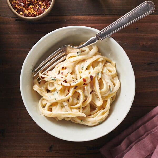 Creamy Fettuccine Alfredo in bowl with red pepper flakes on table.