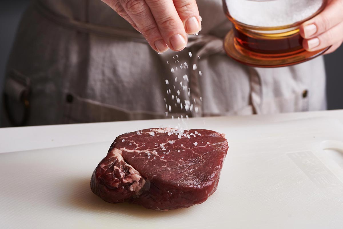 Woman sprinkling salt onto an uncooked filet mignon.