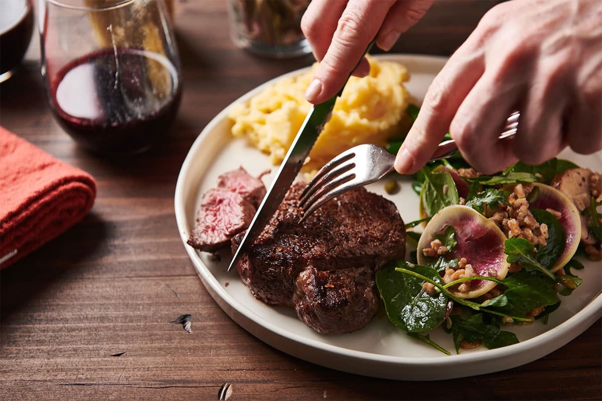 Cutting filet mignon steak on plate with salad and mashed potatoes.