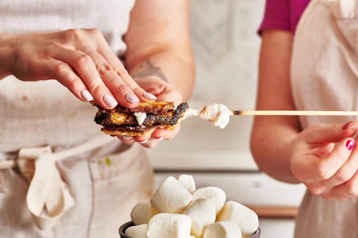 Woman squeezing a marshmallow on a skewer with two pieces of matzoh chocolate.