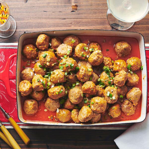 Buffalo chicken meatballs in serving dish on table with red cloth, bowl, and silverware.