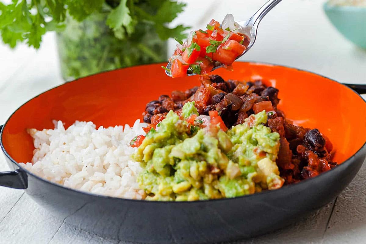 Adding salsa to black beans and rice with guacamole in black and orange bowl.