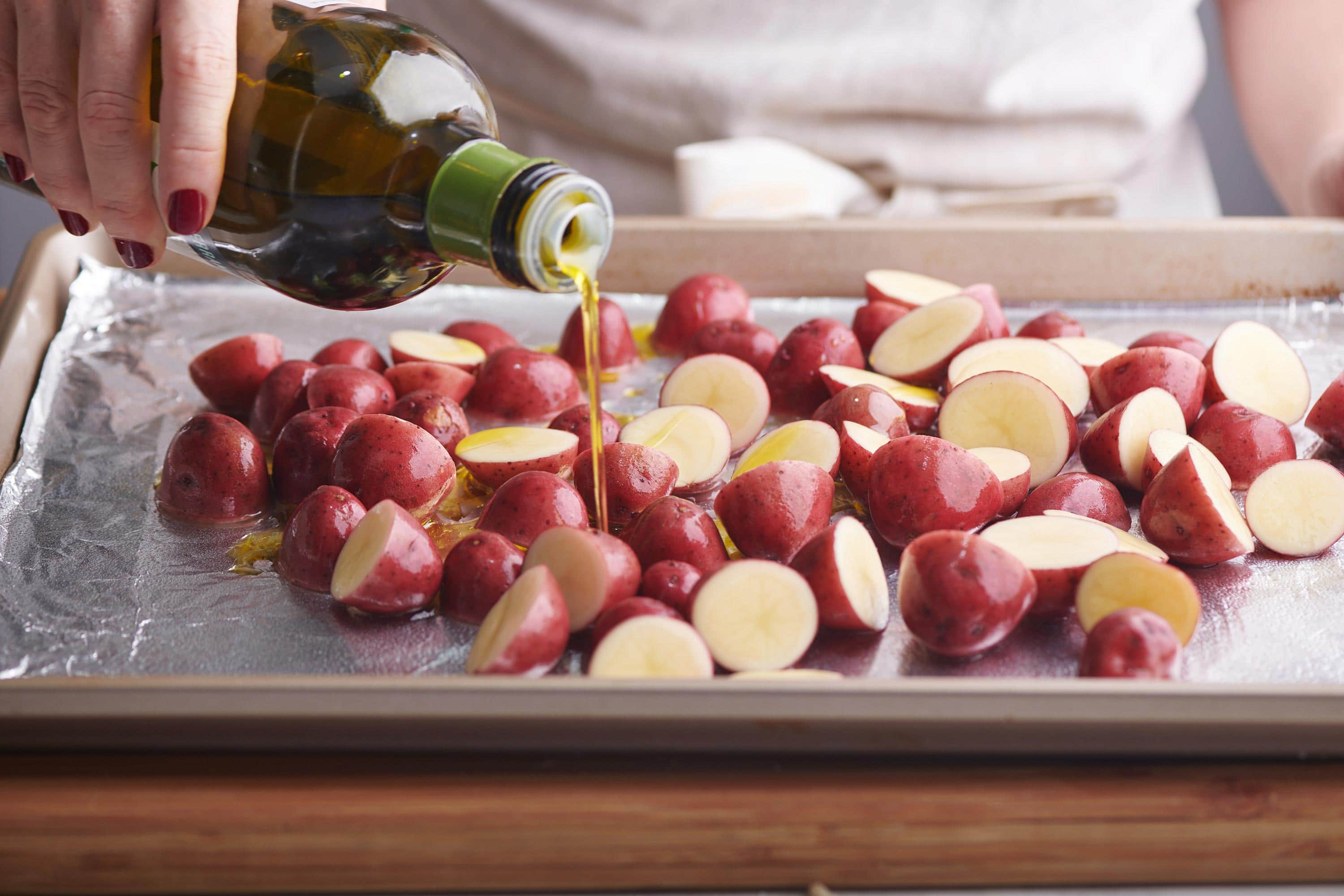 Olive oil drizzling on a foil lined baking sheet of halved red potatoes.