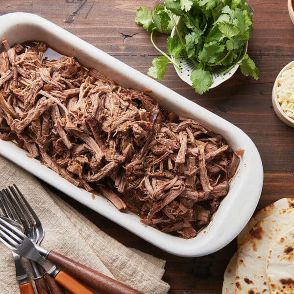 Barbacoa beef in a dish on table with tortillas and taco toppings.