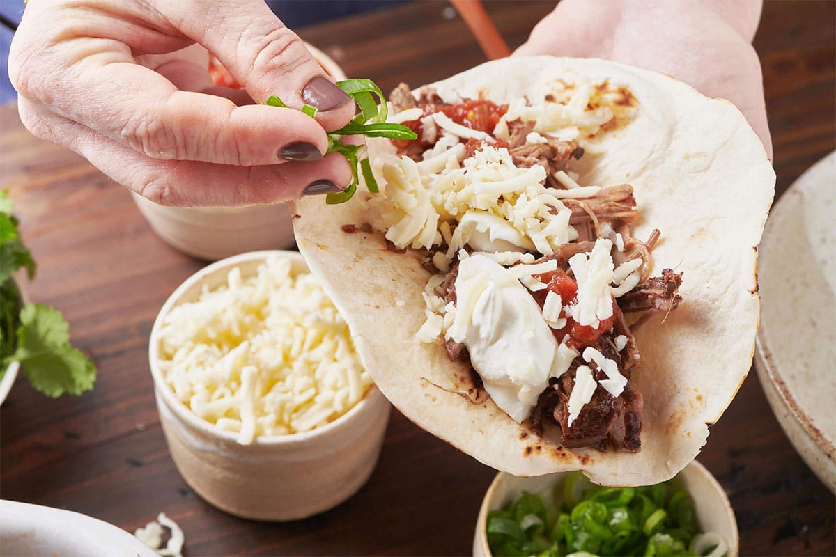 Woman topping barbacoa beef taco in tortilla with shredded cheese, salsa, and sour cream.