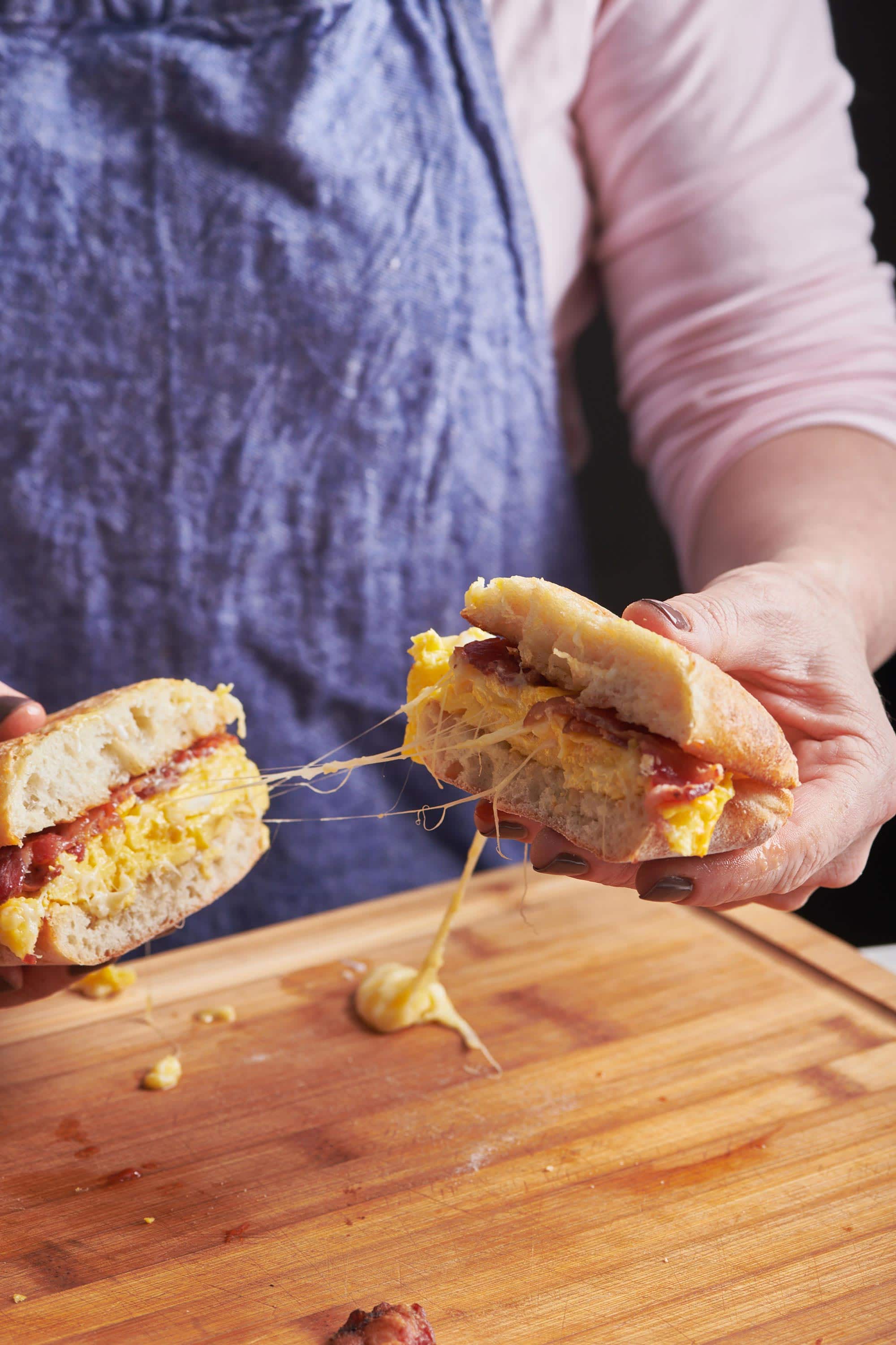 Woman holding two halves of a Classic Bacon, Egg and Cheese Sandwich, dripping with cheese.