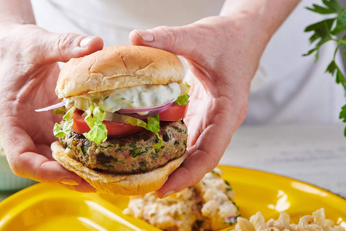 Woman holding turkey burger with tzatziki and other toppings over yellow plate.
