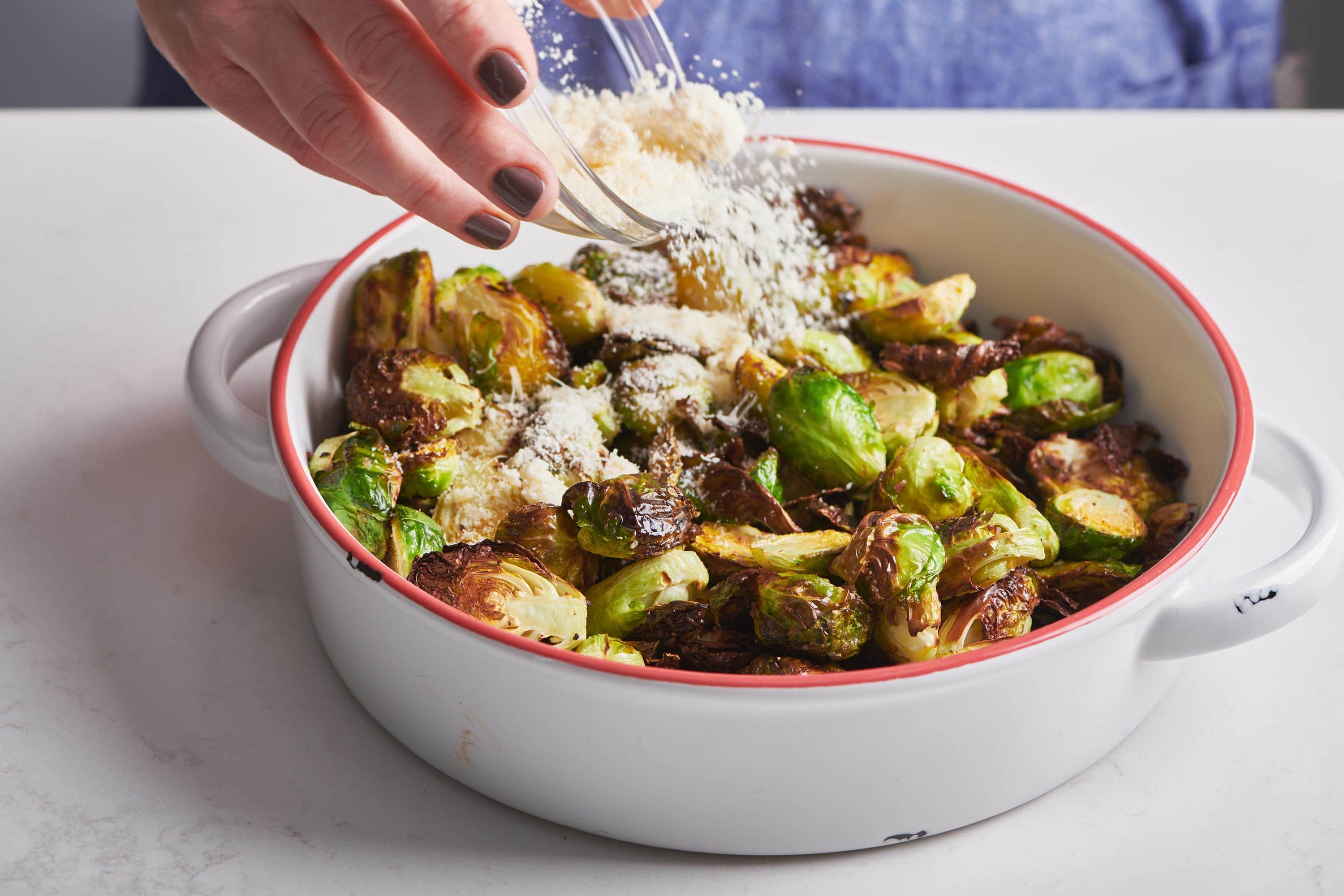 Woman adding grated Parmesan to bowl of air-fried Brussels sprouts.
