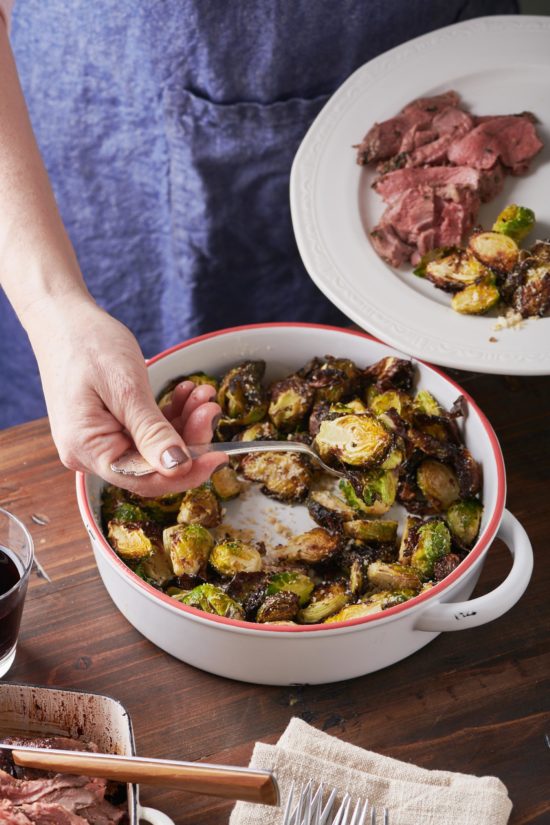 Woman spooning air fryer Brussel sprouts onto a white serving plate with meat.
