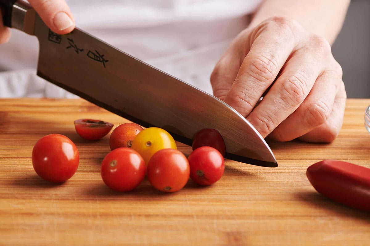 Slicing cherry tomatoes with large chefs knife on wood cutting board.