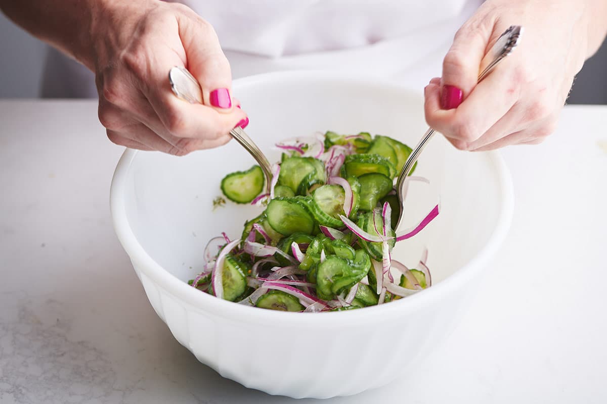 Woman tossing cucumber salad with dressing in white bowl.