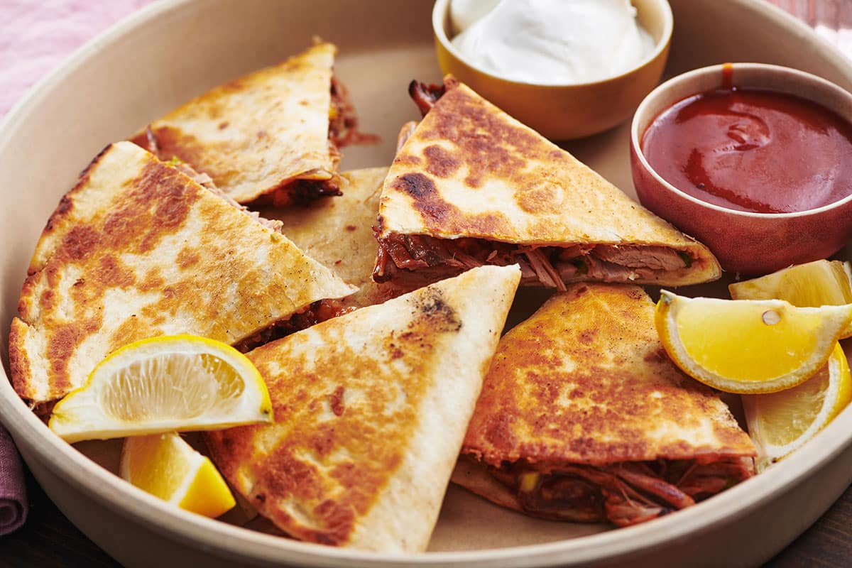Pulled Pork Quesadillas on a tray with barbecue sauce, sour cream, and lemon wedges.