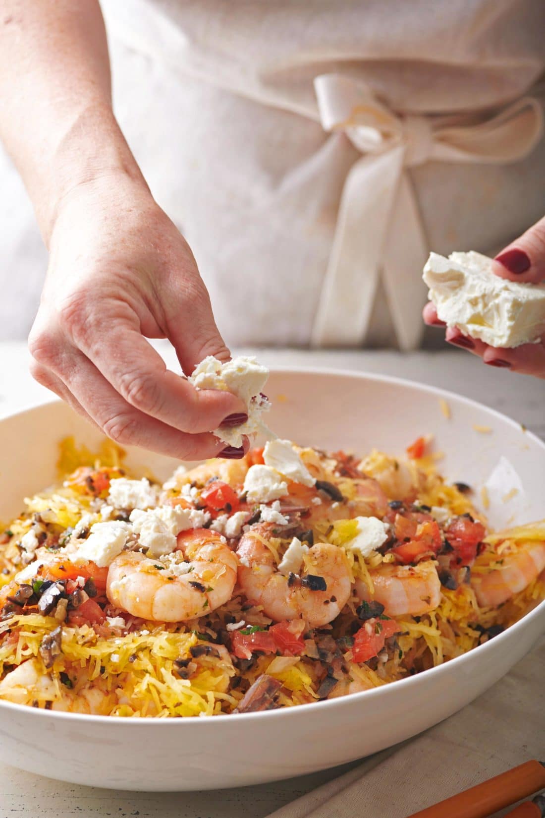 Woman crumbling feta onto a bowl of Greek-Style Spaghetti Squash with Shrimp.