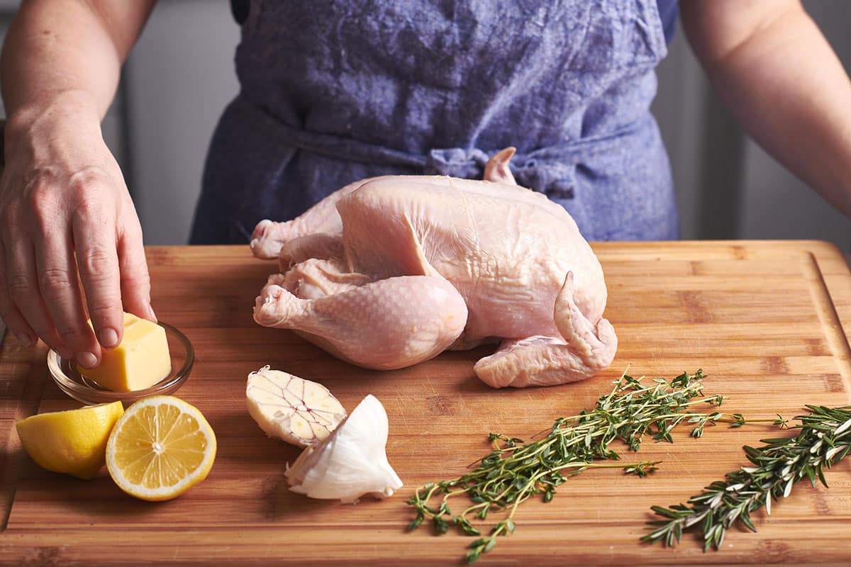 Raw whole chicken on cutting board with lemons, garlic, and herbs.