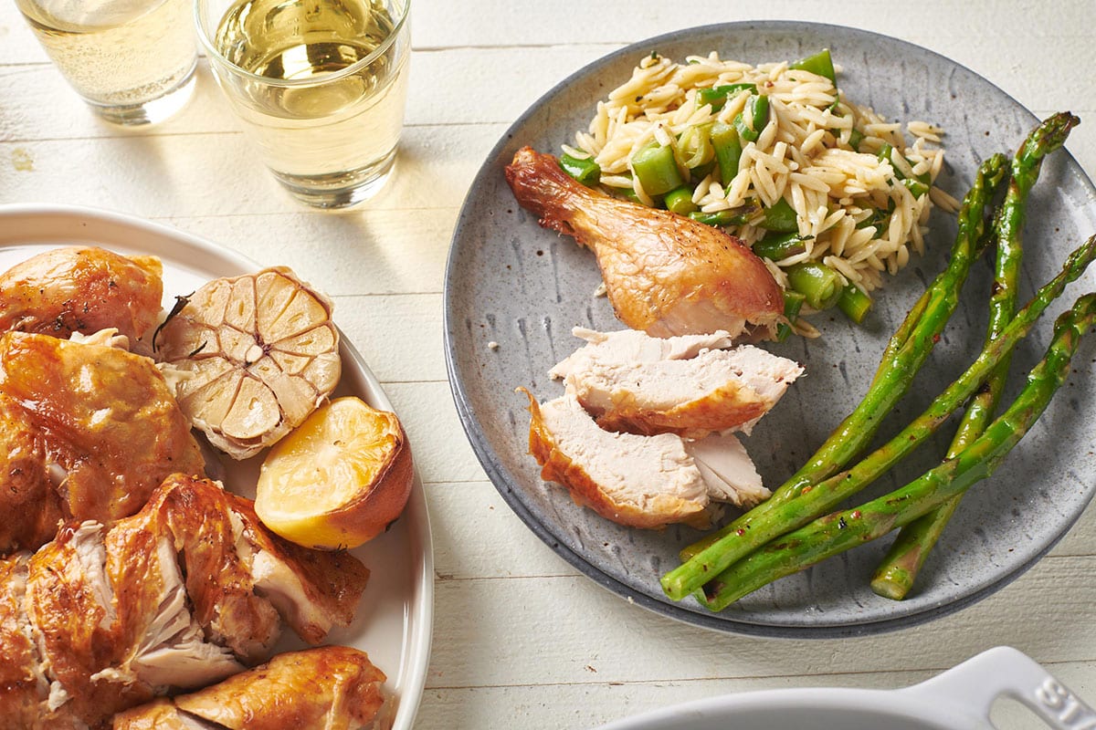 Sliced roast chicken on plates with asparagus, salad, lemon, garlic, and drinks on table.