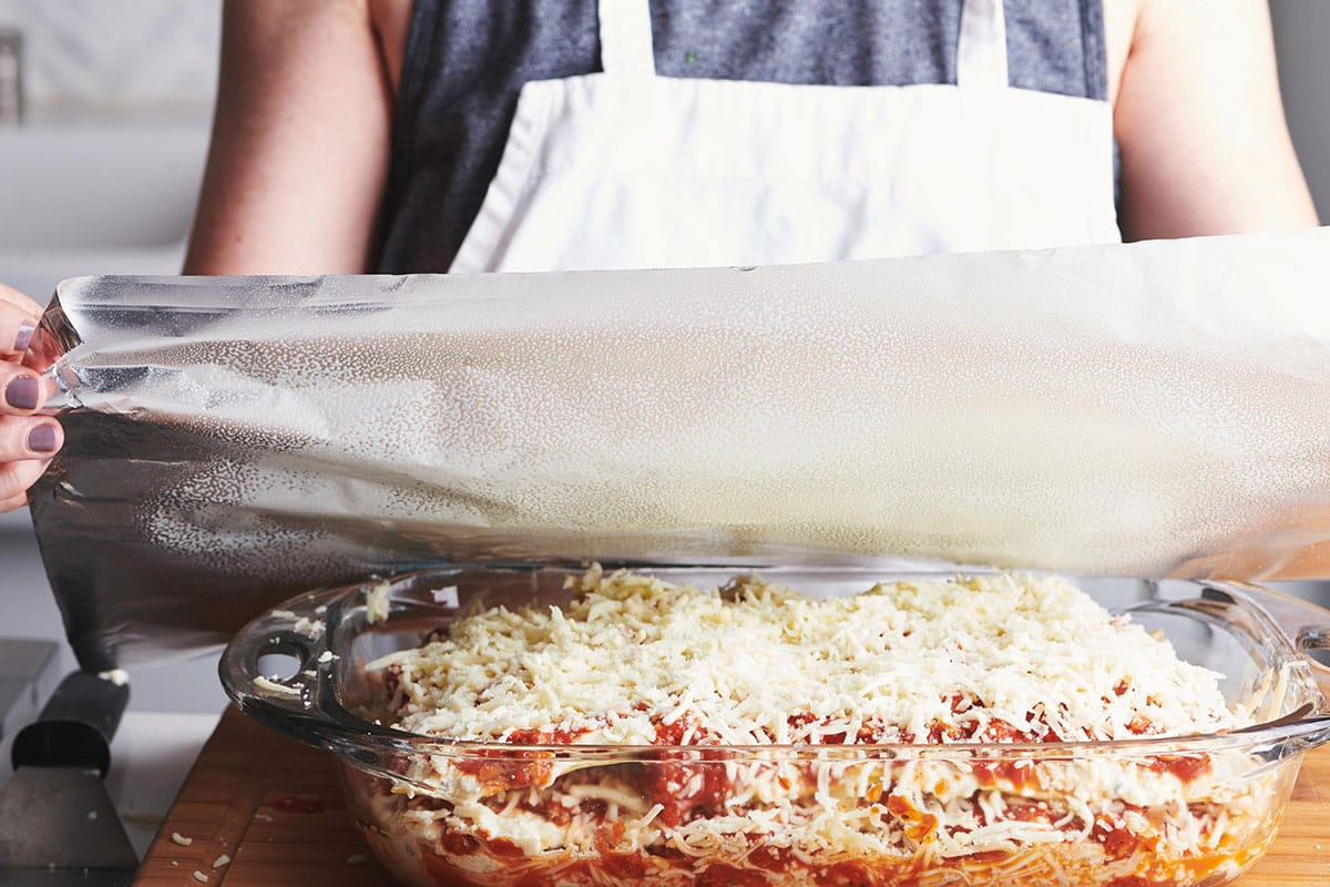 Woman covering unbaked turkey lasagna with oil-sprayed foil.