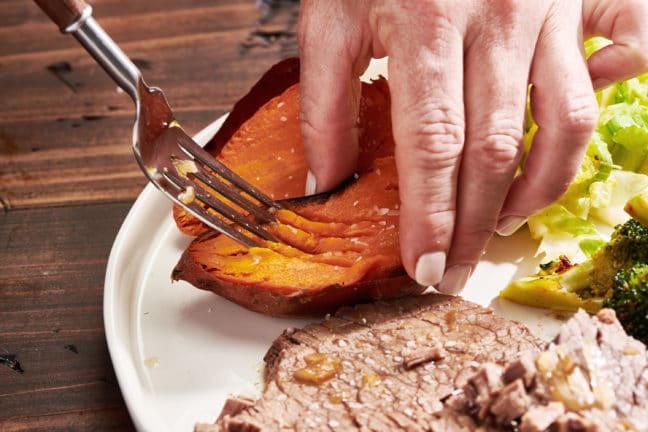Woman using a fork to mash the inside of a baked sweet potato.