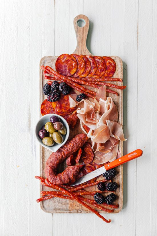 Meat arranged on a board with fruit and a bowl of olives.
